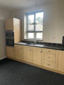 Property for sale by Auction A modern kitchen with light wood cabinets, black countertops, and stainless steel sink beneath a window with white blinds. The left side features built-in appliances, including an oven and a microwave. The flooring is dark-colored. Presented by Palace Auctions