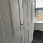 Property for sale by Auction A white wooden built-in wardrobe with paneled doors is situated in a corner of a room with white walls and dark carpet. The wardrobe is adjacent to a window with partially open blinds, allowing some natural light to enter and offering a view of houses and greenery outside. Presented by Palace Auctions