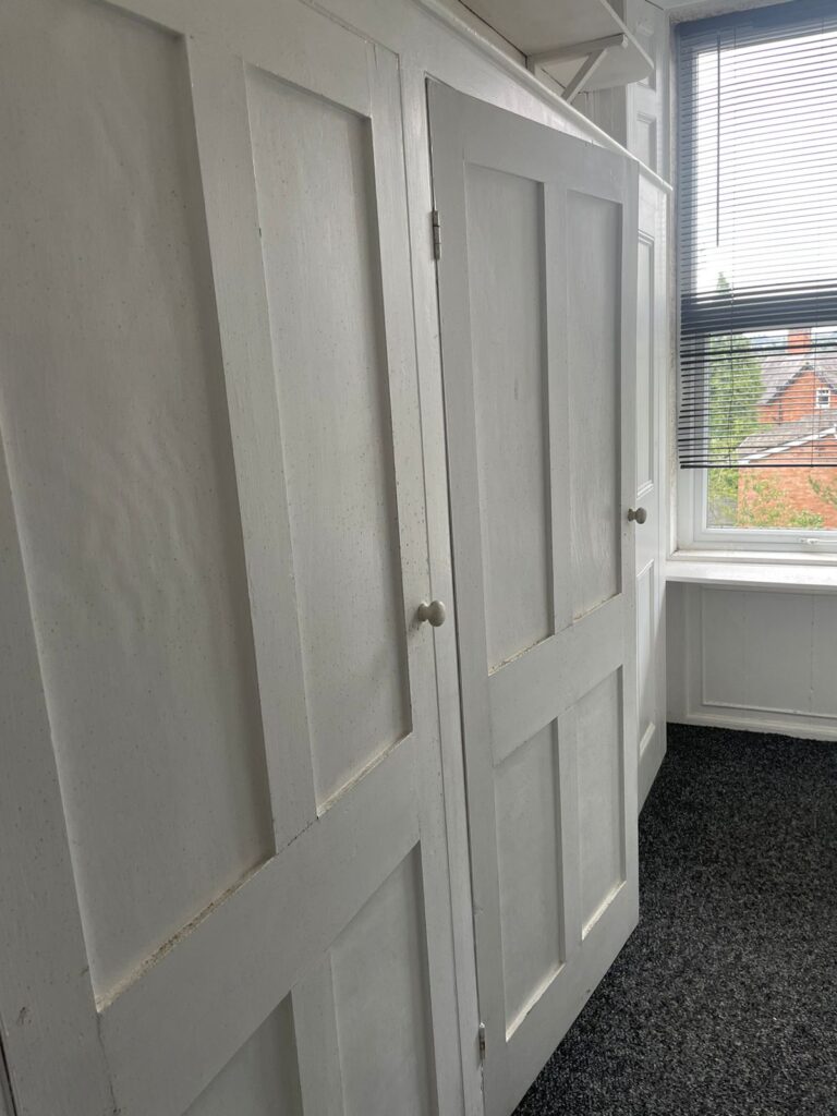 Property for sale by Auction A white wooden built-in wardrobe with paneled doors is situated in a corner of a room with white walls and dark carpet. The wardrobe is adjacent to a window with partially open blinds, allowing some natural light to enter and offering a view of houses and greenery outside. Presented by Palace Auctions