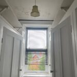 Property for sale by Auction A narrow room with white paneled walls and a central double window with venetian blinds, partially open to reveal an outside view of a red brick building and green foliage. There's a small hanging light fixture on the ceiling. The room contains two open closet doors. Presented by Palace Auctions