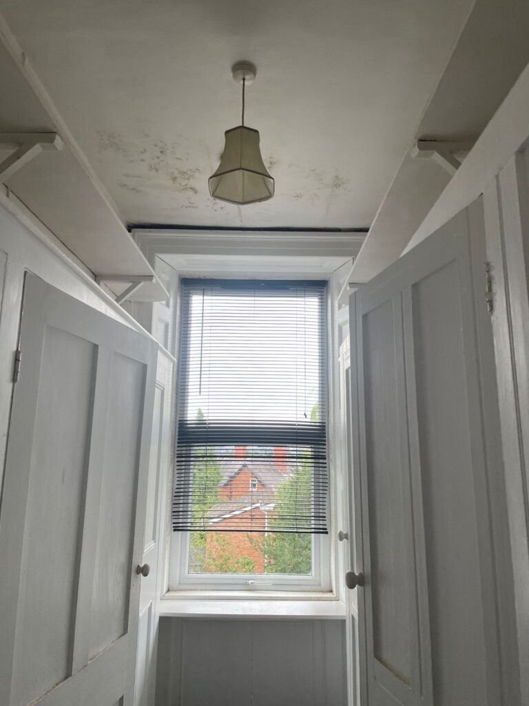 Property for sale by Auction A narrow room with white paneled walls and a central double window with venetian blinds, partially open to reveal an outside view of a red brick building and green foliage. There's a small hanging light fixture on the ceiling. The room contains two open closet doors. Presented by Palace Auctions
