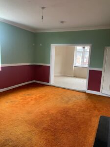 Property for sale by Auction A partially furnished room with green and red walls, an orange carpet, and a white door. An adjacent room with white walls and a beige carpet is visible through a large open doorway. Natural light shines through the windows in both rooms. Presented by Palace Auctions