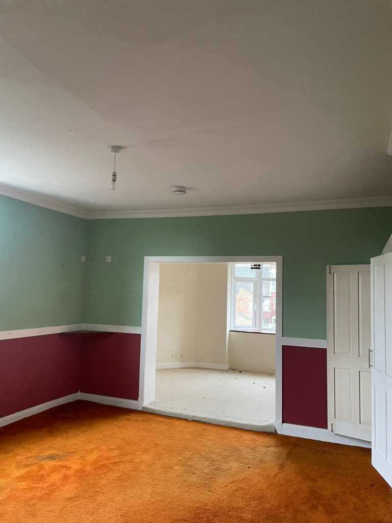 Property for sale by Auction An empty room with green and burgundy walls, white trim, and orange carpet. There is a doorway leading to an adjoining room with white walls and light-colored carpet. A window in the far room allows natural light to enter. Light fixtures hang from the ceilings. Presented by Palace Auctions