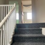 Property for sale by Auction A carpeted staircase with a white railing leads up to an open door revealing a small room. The room appears to have an unfinished ceiling with exposed insulation. Sunlight shines through two windows on the far side of the room. Presented by Palace Auctions