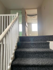 Property for sale by Auction A carpeted staircase with a white railing leads up to an open door revealing a small room. The room appears to have an unfinished ceiling with exposed insulation. Sunlight shines through two windows on the far side of the room. Presented by Palace Auctions