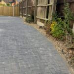 Property for sale by Auction A pathway made of grey pavers curves alongside a tall wooden fence in a residential area. The fence is held up by wooden posts and has some plants and flowers growing at its base. The sky is partly cloudy. Presented by Palace Auctions