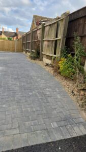 Property for sale by Auction A pathway made of grey pavers curves alongside a tall wooden fence in a residential area. The fence is held up by wooden posts and has some plants and flowers growing at its base. The sky is partly cloudy. Presented by Palace Auctions