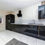 Property for sale by Auction A modern kitchen with glossy black cabinets and gold handles, white tiled backsplash, and white countertops. There's an oven built into the cabinet and a black range hood above the stove. The floor is white tiled, and there's a large window on the left. Presented by Palace Auctions