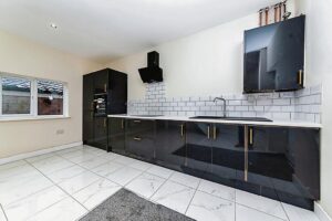 Property for sale by Auction A modern kitchen with glossy black cabinets and gold handles, white tiled backsplash, and white countertops. There's an oven built into the cabinet and a black range hood above the stove. The floor is white tiled, and there's a large window on the left. Presented by Palace Auctions