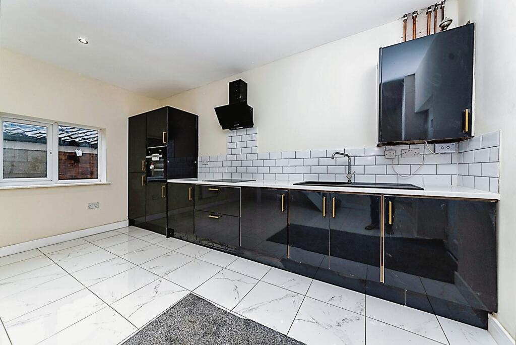 Property for sale by Auction A modern kitchen with glossy black cabinets and gold handles, white tiled backsplash, and white countertops. There's an oven built into the cabinet and a black range hood above the stove. The floor is white tiled, and there's a large window on the left. Presented by Palace Auctions