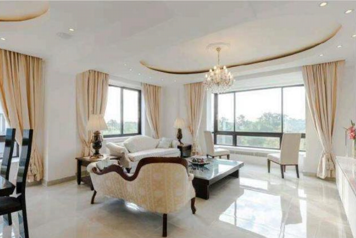 Property for sale by Auction A bright and spacious living room with large windows, cream-colored curtains, and beige furniture, including a sofa and armchairs. A chandelier hangs from the ceiling, and there is a coffee table in the center. Natural light fills the room. Presented by Palace Auctions