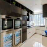 Property for sale by Auction Modern kitchen featuring sleek black and white cabinets, two built-in ovens, a wine cooler stocked with bottled water, and a large window letting in natural light. The countertop includes various kitchen appliances and a decorative blue bowl. Presented by Palace Auctions