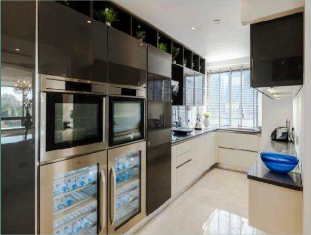 Property for sale by Auction Modern kitchen featuring sleek black and white cabinets, two built-in ovens, a wine cooler stocked with bottled water, and a large window letting in natural light. The countertop includes various kitchen appliances and a decorative blue bowl. Presented by Palace Auctions