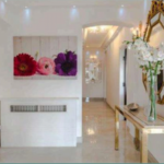Property for sale by Auction A modern hallway with white walls and glossy floor tiles. A large mirror with a decorative gold frame and a slim table holding white flowers adorn the right wall. Across, a radiator covers with a bright floral artwork featuring red, pink, and purple flowers above it. Presented by Palace Auctions