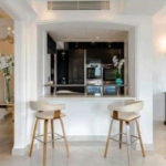 Property for sale by Auction A modern kitchen with a small bar area featuring two stools. The space has white cabinetry, stainless steel appliances, and a glass door refrigerator. The adjacent dining area has a large mirror and distinctive light fixtures. White flowers adorn the spaces. Presented by Palace Auctions