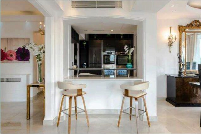 Property for sale by Auction A modern kitchen with a small bar area featuring two stools. The space has white cabinetry, stainless steel appliances, and a glass door refrigerator. The adjacent dining area has a large mirror and distinctive light fixtures. White flowers adorn the spaces. Presented by Palace Auctions