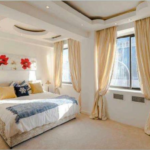 Property for sale by Auction A spacious bedroom with a large bed adorned with white and blue bedding and decorative pillows. The room features two windows with beige curtains, and the walls are decorated with a painting of red flowers. Soft natural light illuminates the room through the windows. Presented by Palace Auctions