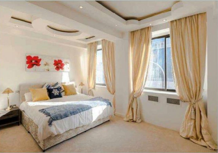 Property for sale by Auction A spacious bedroom with a large bed adorned with white and blue bedding and decorative pillows. The room features two windows with beige curtains, and the walls are decorated with a painting of red flowers. Soft natural light illuminates the room through the windows. Presented by Palace Auctions
