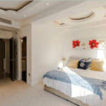 Property for sale by Auction A well-lit modern bedroom with a large bed covered in a white duvet, blue throw, and several pillows. Two bedside tables with lamps are on either side. A decorative headboard with red flowers hangs above the bed. There are two doors leading to other rooms. Presented by Palace Auctions