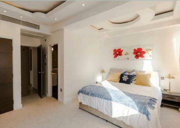 Property for sale by Auction A well-lit modern bedroom with a large bed covered in a white duvet, blue throw, and several pillows. Two bedside tables with lamps are on either side. A decorative headboard with red flowers hangs above the bed. There are two doors leading to other rooms. Presented by Palace Auctions