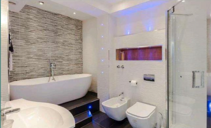 Property for sale by Auction Modern bathroom with a freestanding white bathtub against a tiled wall, a sleek glass-enclosed shower, and a toilet next to a bidet. Ambient blue lighting accents the clean, minimalist design. White fixtures and fittings complement the contemporary aesthetic. Presented by Palace Auctions