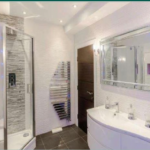 Property for sale by Auction A modern bathroom featuring a glass shower enclosure with a sleek, tile accent wall, a large rectangular mirror above a curved double-sink vanity, a wall-mounted stainless steel towel heater, and contemporary recessed lighting. The room has a minimalist design. Presented by Palace Auctions