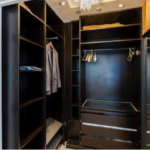 Property for sale by Auction A walk-in closet with dark wooden shelving and drawers. One section has hanging clothes, including a jacket, a shirt, and a few hangers. The other sections have empty shelves and a drawer unit. The closet is well-lit with ceiling lights. Presented by Palace Auctions