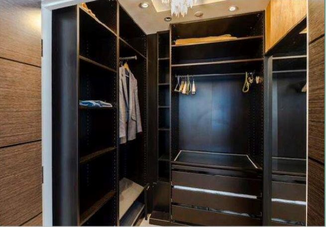 Property for sale by Auction A walk-in closet with dark wooden shelving and drawers. One section has hanging clothes, including a jacket, a shirt, and a few hangers. The other sections have empty shelves and a drawer unit. The closet is well-lit with ceiling lights. Presented by Palace Auctions
