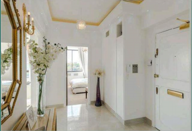 Property for sale by Auction A bright, elegant hallway with white walls and marble flooring. There is a gold-framed mirror and wall sconce on the left wall. The entrance to a bedroom is visible at the end of the hallway. A tall vase with flowers stands near the bedroom entrance. Light streams in from a window in the bedroom. Presented by Palace Auctions