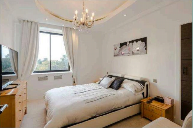Property for sale by Auction A cozy bedroom featuring a double bed with white bedding and several pillows, a wooden dresser with a TV on top, and nightstands on either side of the bed. A large window with white curtains brightens the room. A chandelier hangs from the ceiling. A flower painting is above the headboard. Presented by Palace Auctions