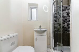 Property for sale by Auction A small bathroom features a compact white toilet, a white vanity with a small sink and single faucet, a narrow frosted window, and a glass-enclosed corner shower with a modern black and white tile pattern. The walls are painted a light color, underscoring the substantial residential and development opportunity. Presented by Palace Auctions