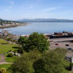 Property for sale by Auction A scenic coastal view of a small town with lush green parks, a pier with an orange-roofed building, and calm blue waters extending to distant mountains under a clear blue sky. Nearby roads and houses line the shoreline, creating a picturesque landscape. Presented by Palace Auctions