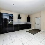 Property for sale by Auction A modern kitchen with glossy black cabinets, white countertops, grey backsplash tiles, and built-in appliances presents a substantial residential and development opportunity. The space features white tiled flooring, recessed lighting, a window on the left, and a door on the right leading to another room. Presented by Palace Auctions