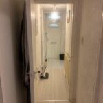 Property for sale by Auction View of a hallway at 94 Mortlake Road, featuring light-colored wooden flooring and a door with decorative glass at the end. A mop and bucket rest on the floor under the glow of a lit ceiling lamp. Coats hang on the left wall, while a radiator is positioned on the right. Presented by Palace Auctions