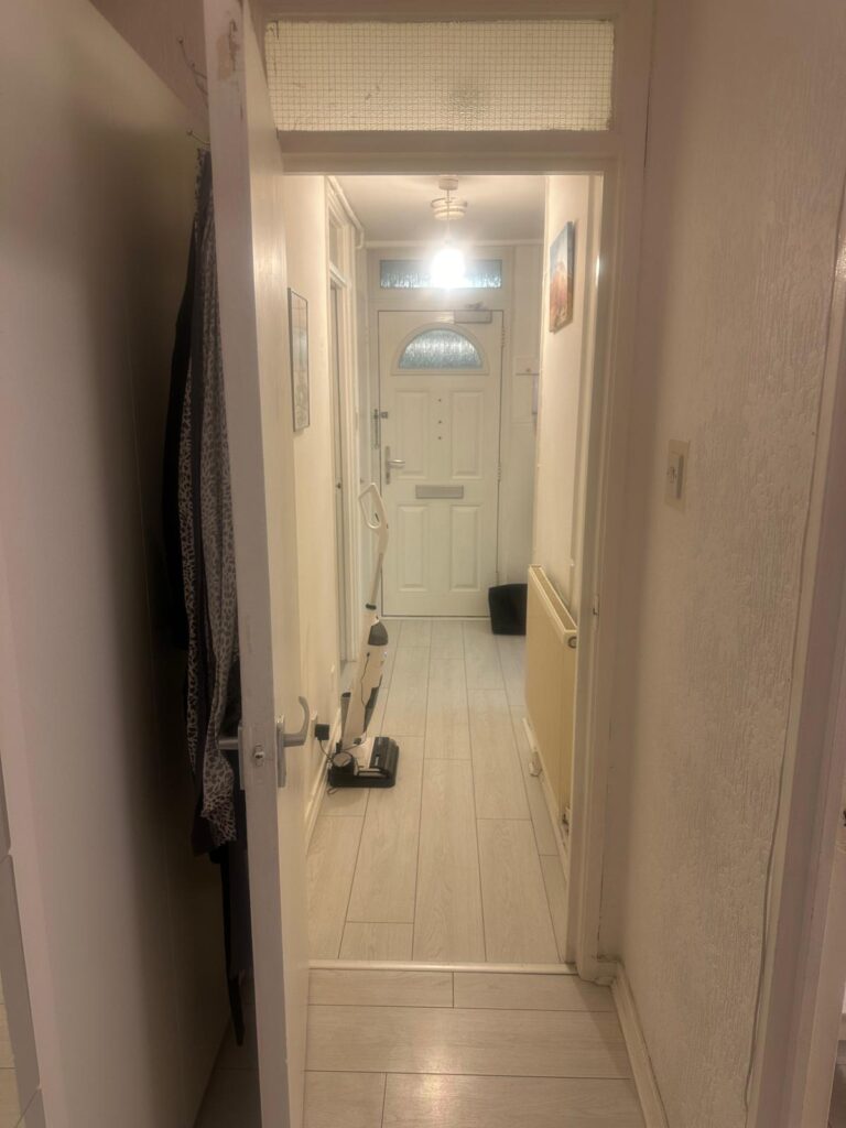 Property for sale by Auction View of a hallway at 94 Mortlake Road, featuring light-colored wooden flooring and a door with decorative glass at the end. A mop and bucket rest on the floor under the glow of a lit ceiling lamp. Coats hang on the left wall, while a radiator is positioned on the right. Presented by Palace Auctions