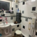 Property for sale by Auction The small bathroom at 94 Mortlake Road boasts white and black tiled walls, a sink adorned with toiletries, a mirror, storage shelves, and a shower with a mounted electric water heater. A gray stool provides seating beneath the high window that illuminates the serene space. Presented by Palace Auctions