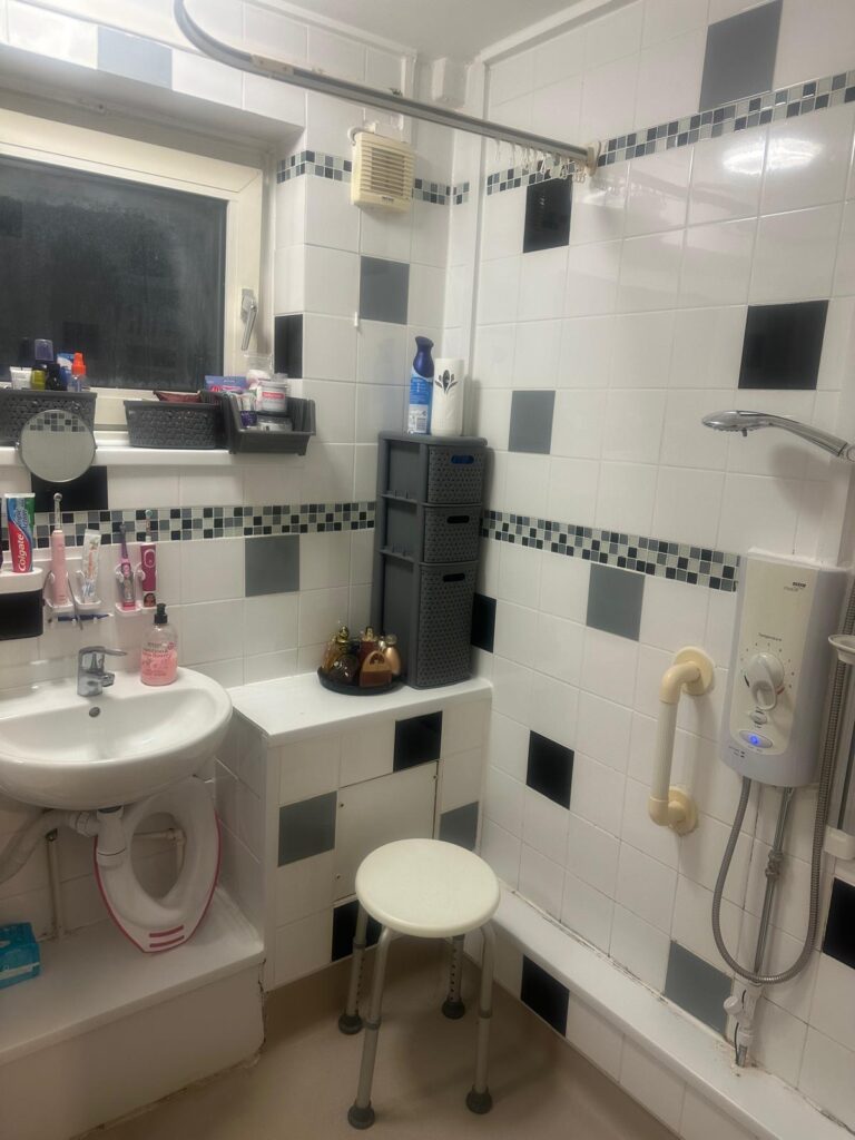 Property for sale by Auction The small bathroom at 94 Mortlake Road boasts white and black tiled walls, a sink adorned with toiletries, a mirror, storage shelves, and a shower with a mounted electric water heater. A gray stool provides seating beneath the high window that illuminates the serene space. Presented by Palace Auctions