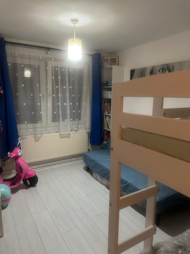 Property for sale by Auction A cozy children's room at 94 Mortlake Road features a bunk bed, blue curtains, and a white polka dot sheer curtain covering the window. There's a pink scooter and a bookshelf brimming with books beside a radiator. Neutral-colored walls and light wood flooring complete this charming space. Presented by Palace Auctions