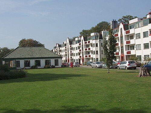 Property for sale by Auction A row of white apartment buildings with balconies stands in the background, with a small white building and a green roof nearby. Several cars are parked along the driveway, and a well-maintained lawn occupies the foreground under a clear blue sky. Presented by Palace Auctions