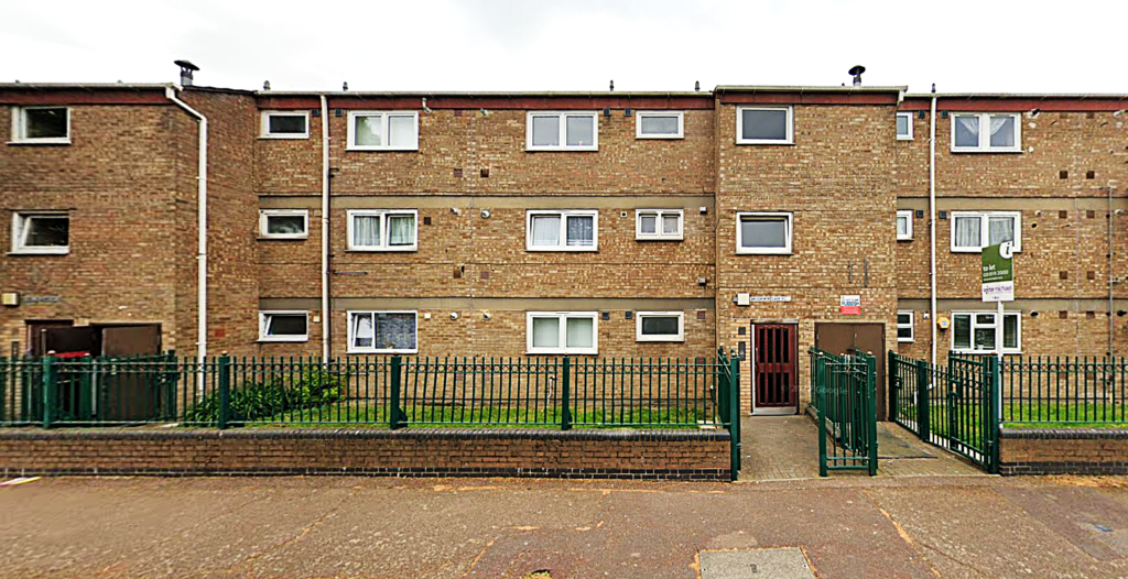 Property for sale by Auction The three-story brick apartment building at 94 Mortlake Road features uniform rectangular windows, bordered by a green metal fence. A small green yard is visible in front, with a sign near the entrance gate. The sky is overcast. Presented by Palace Auctions