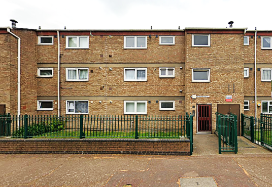 Property for sale by Auction The three-story brick apartment building at 94 Mortlake Road features uniform rectangular windows, bordered by a green metal fence. A small green yard is visible in front, with a sign near the entrance gate. The sky is overcast. Presented by Palace Auctions