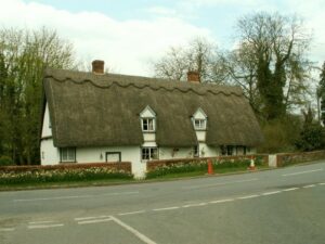 Property for sale by Auction A traditional English thatched cottage with two chimneys and small dormer windows sits by a roadside. Surrounded by trees, a brick wall, and orange traffic cones, it embodies charm. For valuation and marketing advice, consult Palace Auctions under the partly cloudy sky. Presented by Palace Auctions