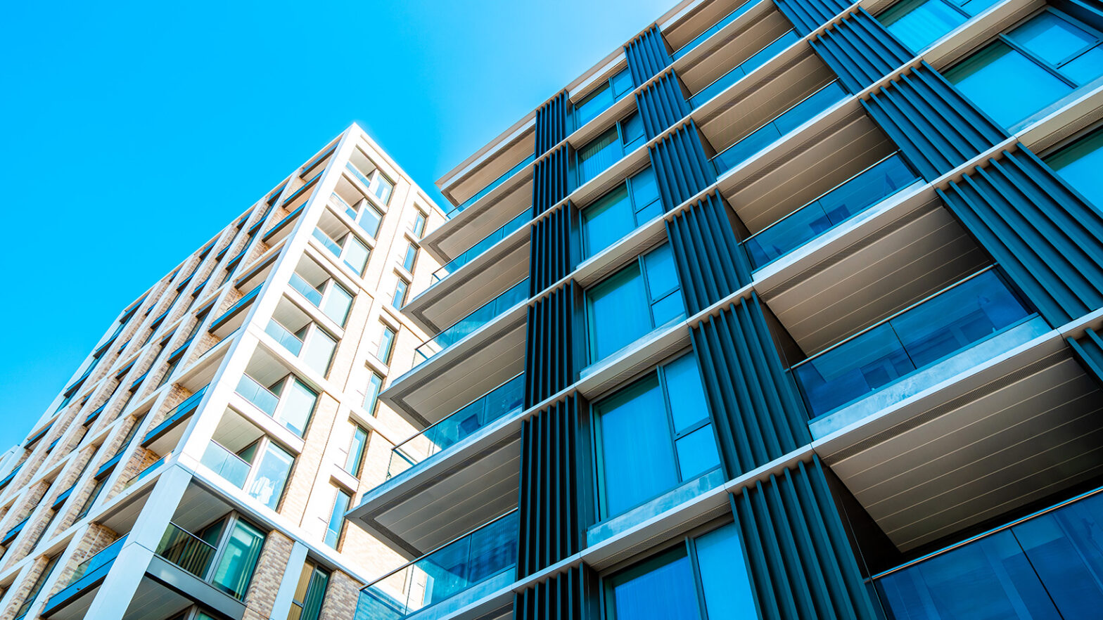 Property for sale by Auction Modern apartment buildings with large glass windows and balconies, viewed from a low angle against a clear blue sky. The structures feature a mix of light-colored bricks and dark vertical paneling, creating a striking architectural contrast. Presented by Palace Auctions