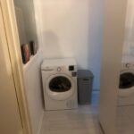 Property for sale by Auction The compact laundry area at 94 Mortlake Road features a white washing machine beside a tall, gray laundry basket. It boasts light-colored wooden flooring and a mirrored wardrobe door that elegantly reflects the room's interior elements. Presented by Palace Auctions