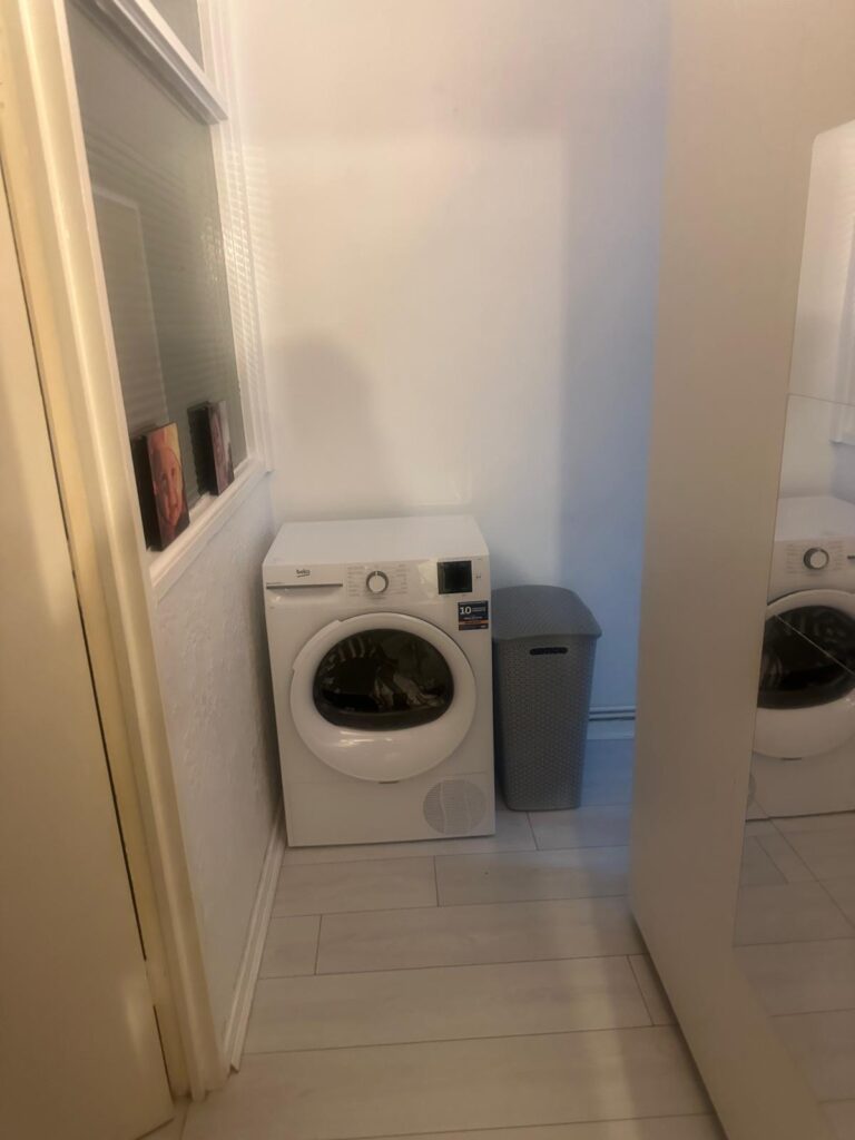 Property for sale by Auction The compact laundry area at 94 Mortlake Road features a white washing machine beside a tall, gray laundry basket. It boasts light-colored wooden flooring and a mirrored wardrobe door that elegantly reflects the room's interior elements. Presented by Palace Auctions