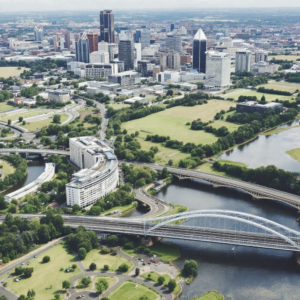 Property for sale by Auction Aerial view of a cityscape in Birmingham, showcasing modern skyscrapers and vast green spaces. Two bridges span a river, surrounded by lush parks. Roads curve through the landscape, with a large unique-shaped building standing out—a promising spot for property investment enthusiasts. Presented by Palace Auctions