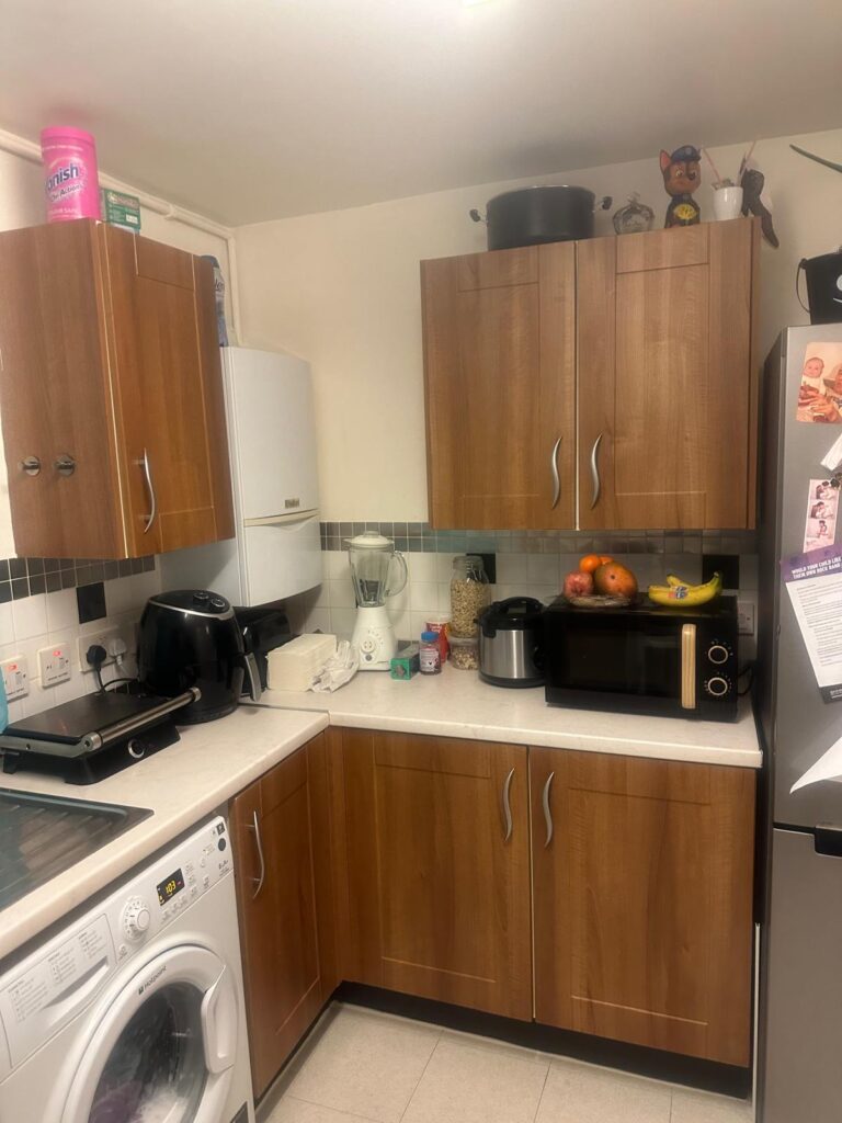 Property for sale by Auction Located at 94 Mortlake Road, this compact kitchen features wooden cabinets. The countertops host a toaster, blender, fruit bowl, rice cooker, and microwave. A washing machine nests below while a small figure decorates a top cabinet. To the right stands a fridge adorned with papers and magnets. Presented by Palace Auctions
