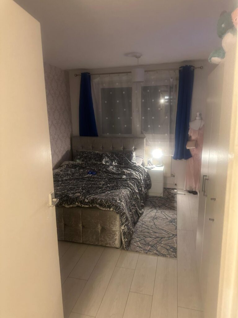 Property for sale by Auction A cozy bedroom at 94 Mortlake Road features a double bed draped with a patterned duvet. The room boasts a window with sheer curtains and dark blue drapes. A bedside lamp glows softly on a small table, while a mannequin adorned in pink stands elegantly in the corner. Presented by Palace Auctions
