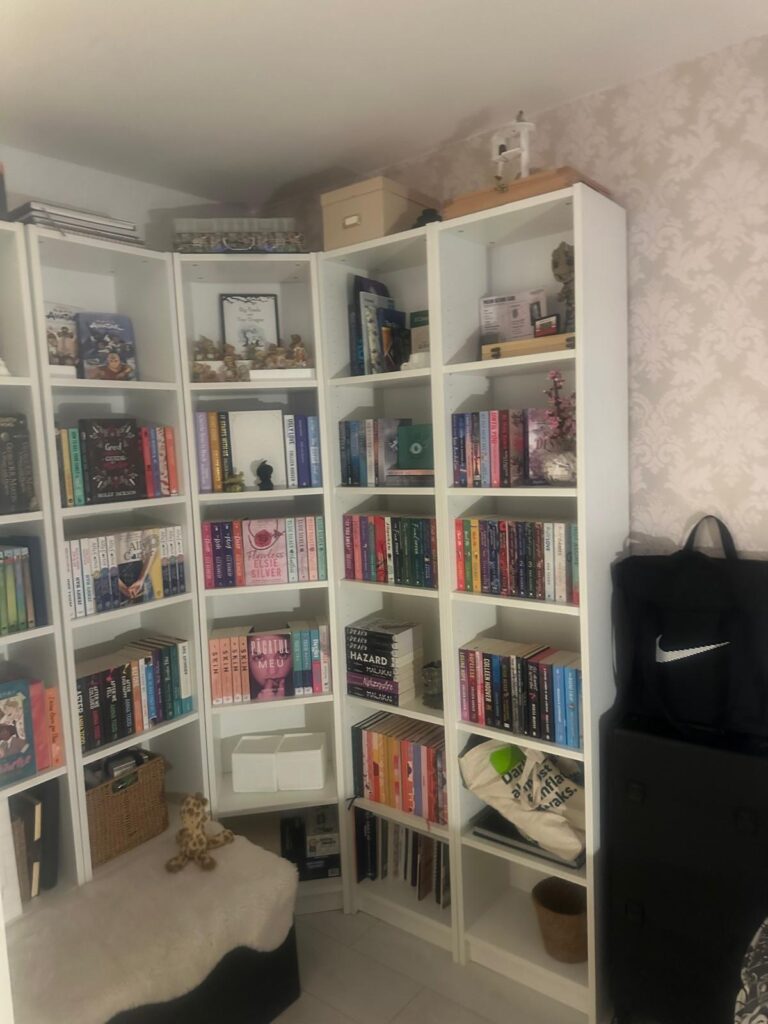 Property for sale by Auction A cozy corner at 94 Mortlake Road features white bookshelves filled with colorful books and decorative items. A small stuffed giraffe rests on a wicker basket, while a large black bag leans against the shelves. The walls boast a subtle, patterned wallpaper, adding charm to the space. Presented by Palace Auctions