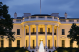 Property for sale by Auction As dusk falls over the White House, lights illuminate the iconic building while an American flag proudly flutters above. A water fountain graces the foreground, framed by lush flowers and trees against a cloudy sky—whispers of Trump's return stirring potential ripples in auction and real estate markets linger. Presented by Palace Auctions